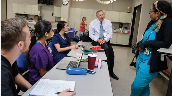Dr. David Brown sits on desk while he speaks to three students in a classroom.