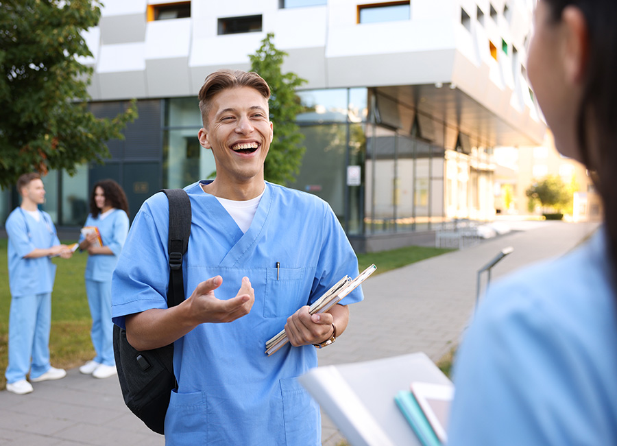 A student in blue scrubs talks with a classmate outside a university building.