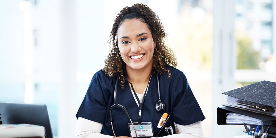 A healthcare professional in scrubs sits at a desk with a laptop and medical reference materials.